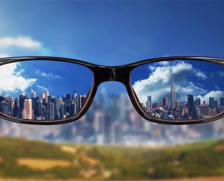 first person view staring through eye glasses at a cityscape. The image inside of the lenses is crystal clear, and the surrounding area is very blurry. This image illustrates myopia (nearsightedness), which is characterized by the inability to see clearly from a distance.